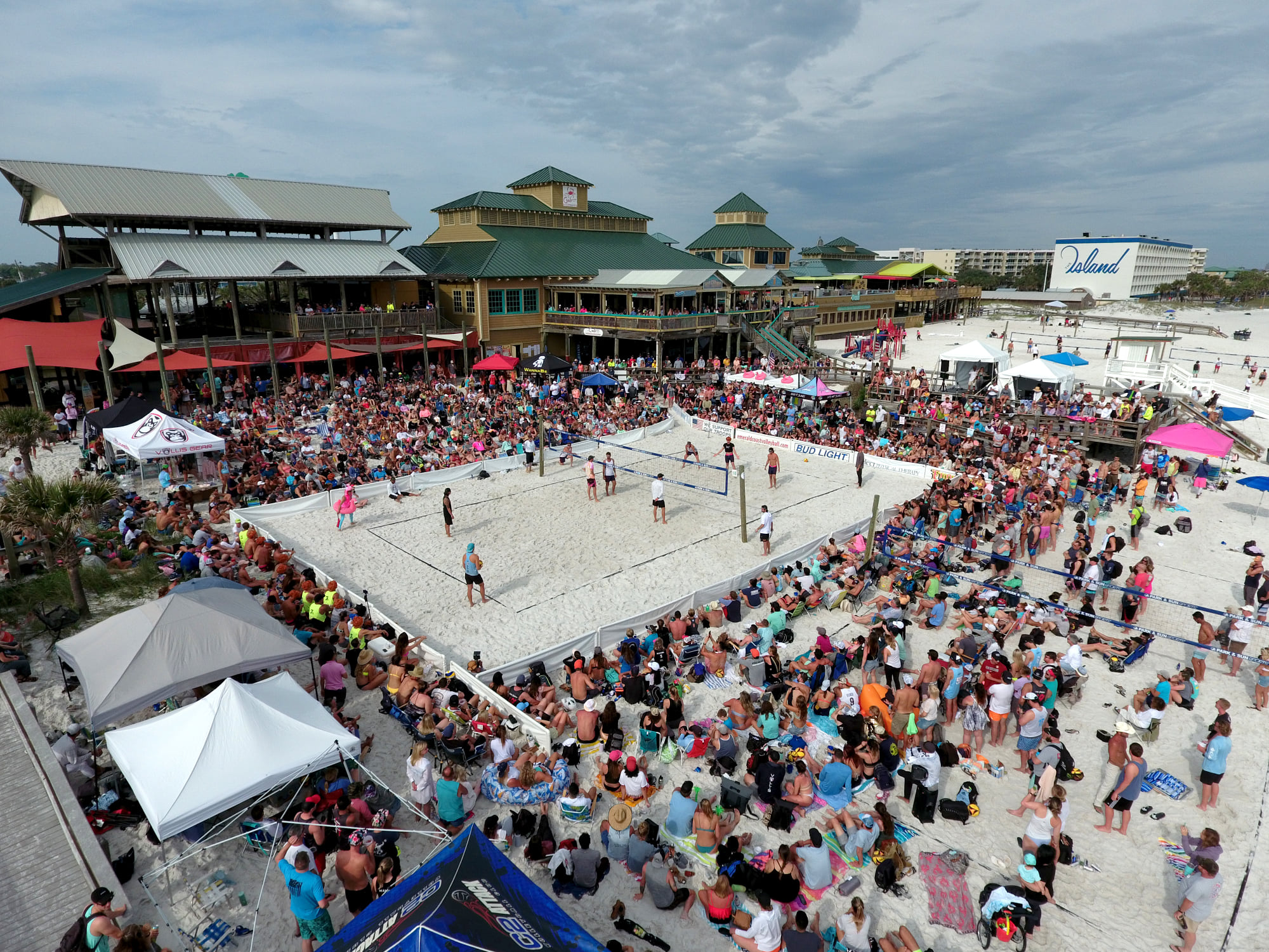 Aerial view of Emerald Coast Volleyball tournament showing multiple beach volleyball courts, thousands of spectators, sponsor banners, and the vibrant carnival atmosphere along the Gulf of Mexico